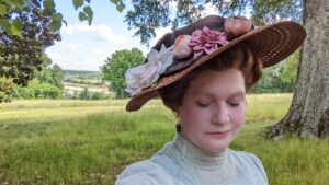 auburn custom wig dressed in an edwardian style updo, topped with a large hat with flowers adorning it.