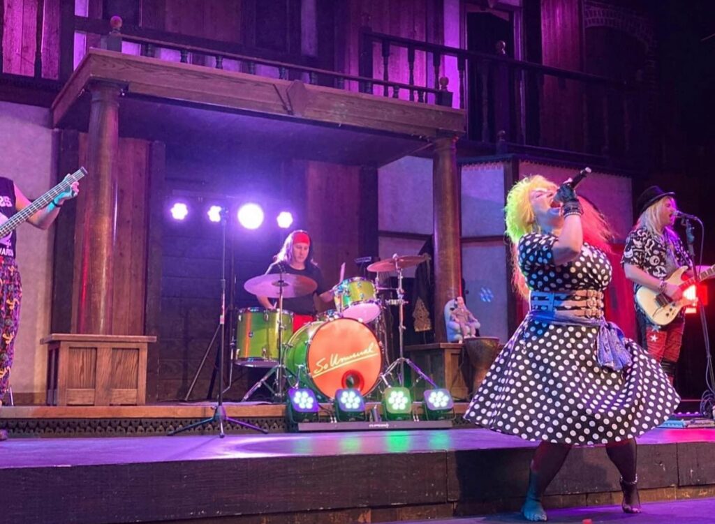 Cyndee Lauper tribute artist Becky from She's So Unusual, performing onstage in a custom red and yellow wig and black and white polka dot dress. The drummer and guitar player are just behind her, the stage is lit in purple