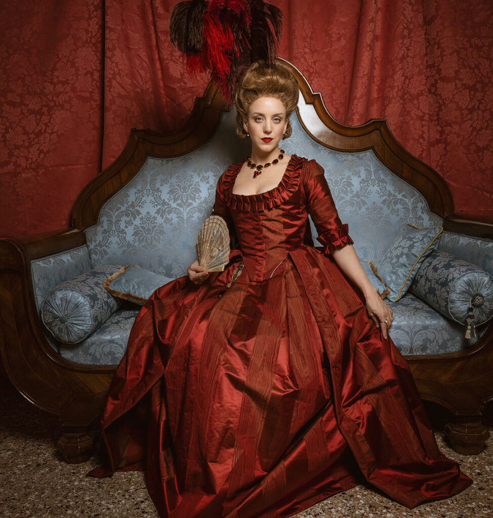 a woman in a red 18th century gown sits on a blue sofa, she's wearing a custom historical wig styled in an 18th century style